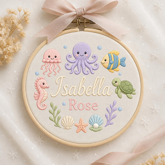 May include: A circular wooden embroidery hoop featuring a light-colored fabric background with embroidered sea creatures including a pink jellyfish, purple octopus, yellow fish, pink seahorse, and green turtle. The text Isabella Rose is centered.