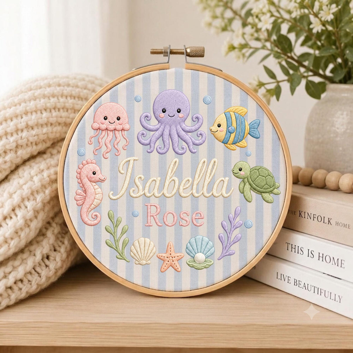 May include: A circular wooden embroidery hoop containing fabric with light blue and white vertical stripes. The fabric features embroidered sea creatures including a purple octopus, pink jellyfish, seahorse, green turtle, and fish, with the name Isabella Rose.