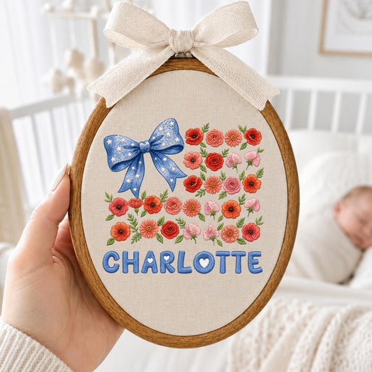 May include: An oval wooden embroidery hoop containing a beige fabric panel. The design features a blue bow with white stars, a cluster of red, pink, and orange flowers, and the name CHARLOTTE in blue letters. A beige fabric bow is tied at the top.