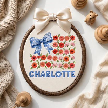 May include: An oval embroidery hoop featuring a cream fabric background with a blue bow with white stars, a collection of red, pink, and orange flowers, and the name CHARLOTTE embroidered in blue letters. A large cream satin bow sits at the top.