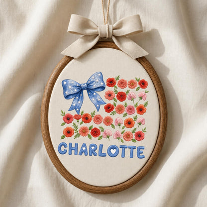 May include: An oval embroidery hoop containing cream fabric with a blue bow featuring white stars, a cluster of red, pink, and orange flowers, and the name CHARLOTTE embroidered in blue letters. A beige ribbon bow is tied at the top of the hoop.