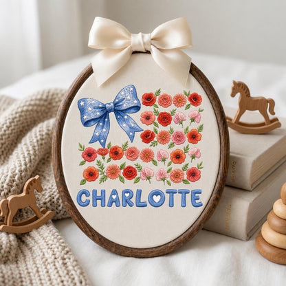 May include: An oval wooden embroidery hoop containing cream fabric with a blue bow featuring white stars, a collection of red, pink, and orange floral embroidery, and the name CHARLOTTE in blue letters. A cream satin ribbon bow is tied at the top.