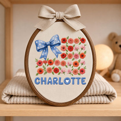 May include: An oval embroidery hoop containing cream fabric with a blue bow featuring white stars, a floral pattern of red and pink flowers with green leaves, and the name CHARLOTTE in blue letters, topped with a beige fabric bow.