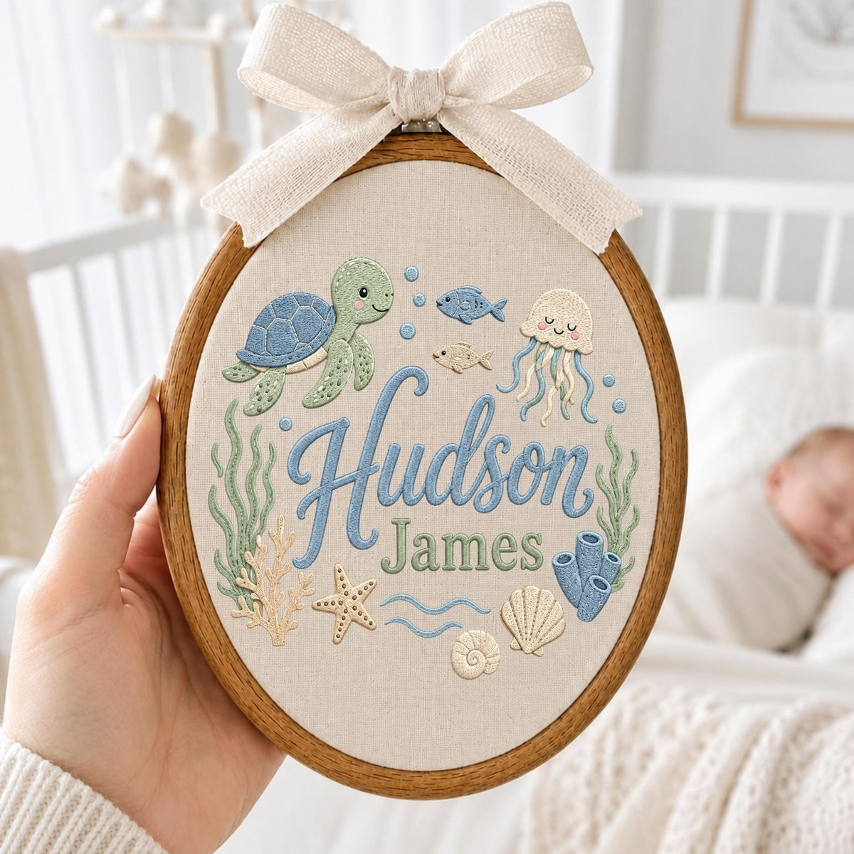 May include: An oval wooden embroidery hoop containing fabric with a stitched sea life design featuring a turtle, jellyfish, fish, coral, and shells. The text Hudson James is embroidered in blue and green. A beige fabric bow is attached to the top.