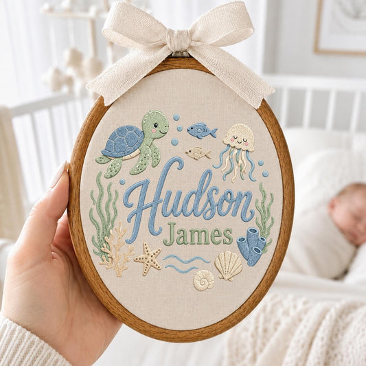 May include: An oval wooden embroidery hoop containing fabric with a stitched sea life design featuring a turtle, jellyfish, fish, coral, and shells. The text Hudson James is embroidered in blue and green. A beige fabric bow is attached to the top.