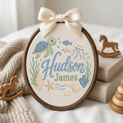 May include: An oval wooden embroidery hoop containing cream fabric with embroidered sea life including a green turtle, jellyfish, starfish, and shells. The name Hudson James is stitched in blue and green script. A cream satin bow tops the hoop.
