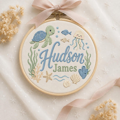 May include: A circular wooden embroidery hoop containing white fabric with the embroidered names Hudson James in blue and green script. The design features a sea turtle, jellyfish, fish, starfish, seashells, coral, and seaweed in soft pastel tones.