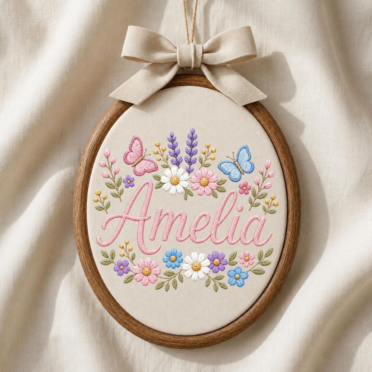 May include: An oval wooden embroidery hoop containing cream fabric with the name Amelia stitched in pink cursive script. The text is surrounded by embroidered flowers in shades of pink, blue, white, and purple, two butterflies, and a beige fabric bow.