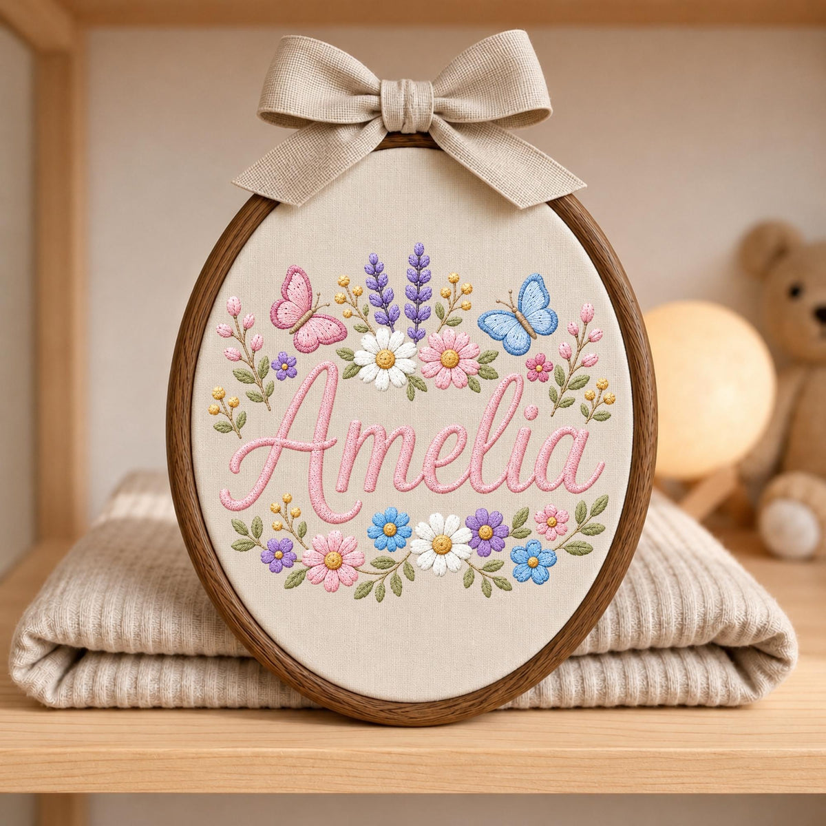 May include: An oval wooden embroidery hoop containing cream fabric with the name Amelia stitched in pink cursive. The design features embroidered pink and blue butterflies, purple lavender sprigs, and various colorful flowers. A beige bow sits on top.