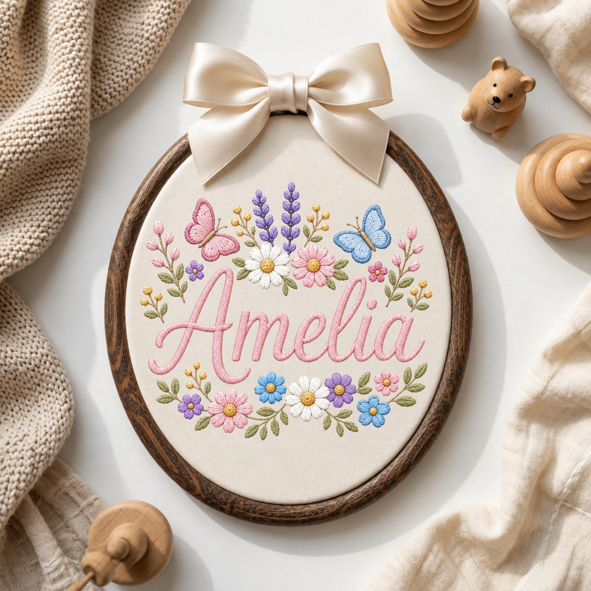 May include: An oval embroidery hoop with a dark wood frame containing cream fabric. The fabric features the name Amelia embroidered in pink script, surrounded by colorful floral motifs, green leaves, and two butterflies, topped with a cream satin bow.