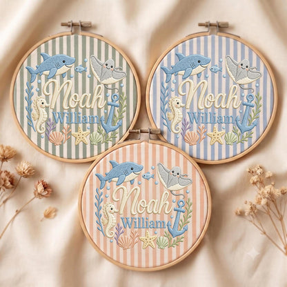 May include: Three circular wooden embroidery hoops featuring striped fabric backgrounds in sage green, light blue, and peach. Each hoop displays embroidered sea life including a shark, stingray, seahorse, anchor, and shells, with the name Noah William.