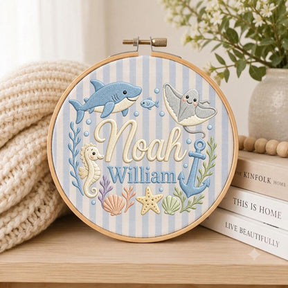 May include: A circular wooden embroidery hoop containing fabric with light blue and white vertical stripes. The embroidery features a blue shark, a gray stingray, a seahorse, a blue anchor, a starfish, and seashells, with the names Noah William.