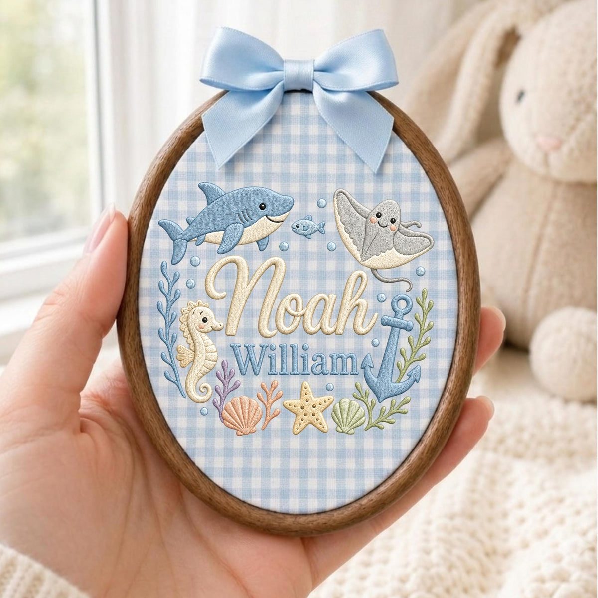 May include: An oval wooden embroidery hoop featuring a light blue gingham fabric background. The embroidery displays the names Noah and William in script and block lettering, surrounded by a shark, stingray, seahorse, anchor, and starfish.