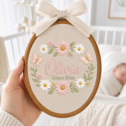 May include: An oval wooden embroidery hoop containing beige fabric with floral embroidery. The design features pink and white daisies, green leaves, and two pink butterflies. The name Olivia Grace Elise is embroidered in pink and green script.