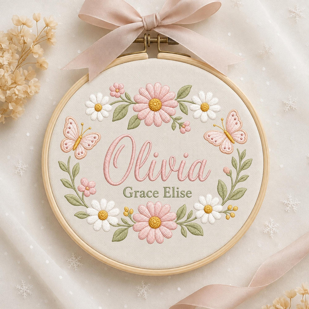 May include: A circular wooden embroidery hoop containing cream fabric with a floral wreath design. The embroidery features pink and white daisies, green leaves, and two pink butterflies. The name Olivia Grace Elise is stitched in pink and green.