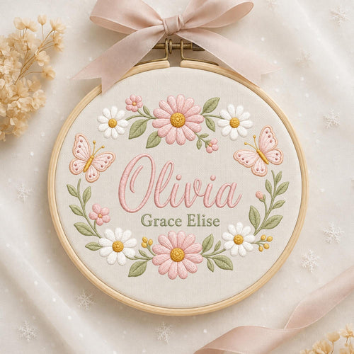 May include: A circular wooden embroidery hoop containing cream fabric with a floral wreath design. The embroidery features pink and white daisies, green leaves, and two pink butterflies. The name Olivia Grace Elise is stitched in pink and green.