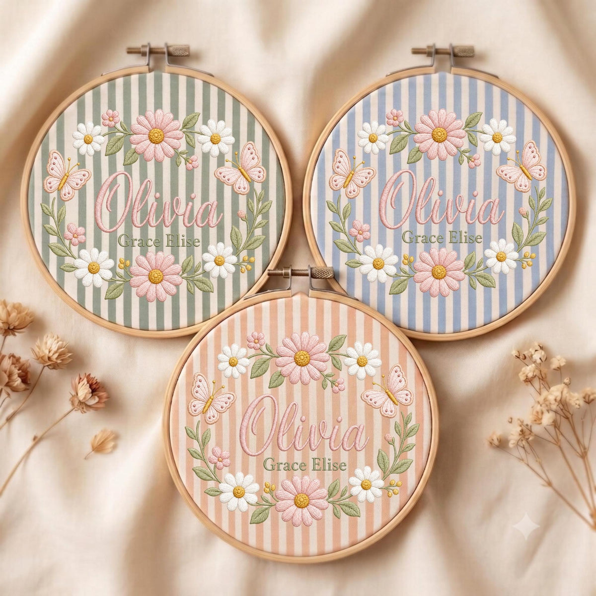 May include: Three circular wooden embroidery hoops featuring striped fabric backgrounds in sage green, light blue, and peach. Each hoop displays embroidered pink and white daisies, butterflies, and the name Olivia Grace Elise in pink script.