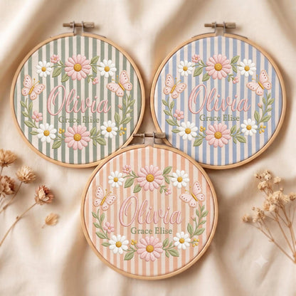 May include: Three circular wooden embroidery hoops featuring striped fabric backgrounds in sage green, light blue, and peach. Each hoop displays embroidered pink and white daisies, butterflies, and the name Olivia Grace Elise in pink script.