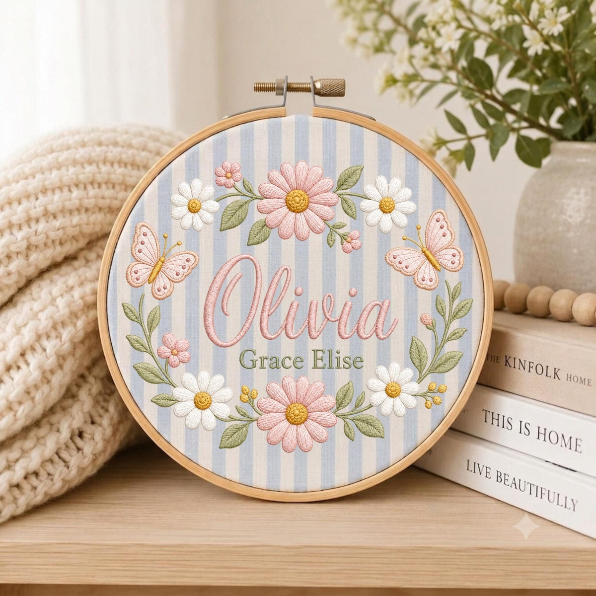 May include: A circular wooden embroidery hoop containing fabric with light blue and white vertical stripes. The embroidery features the name Olivia Grace Elise in pink and green text, surrounded by a wreath of pink and white daisies and butterflies.