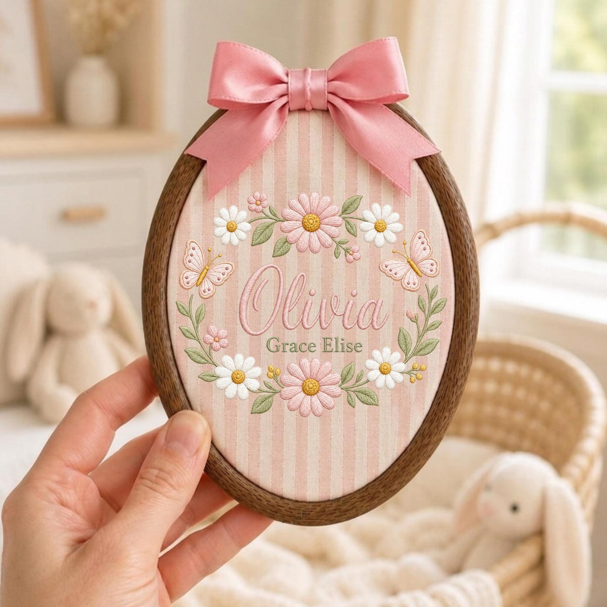 May include: An oval wooden embroidery hoop containing fabric with pink and white vertical stripes. The fabric features embroidered daisies, green leaves, two butterflies, and the text Olivia Grace Elise. A pink satin ribbon bow is attached.