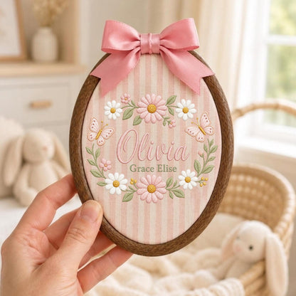 May include: An oval wooden embroidery hoop containing fabric with pink and white vertical stripes. The fabric features embroidered daisies, green leaves, two butterflies, and the text Olivia Grace Elise. A pink satin ribbon bow is attached.