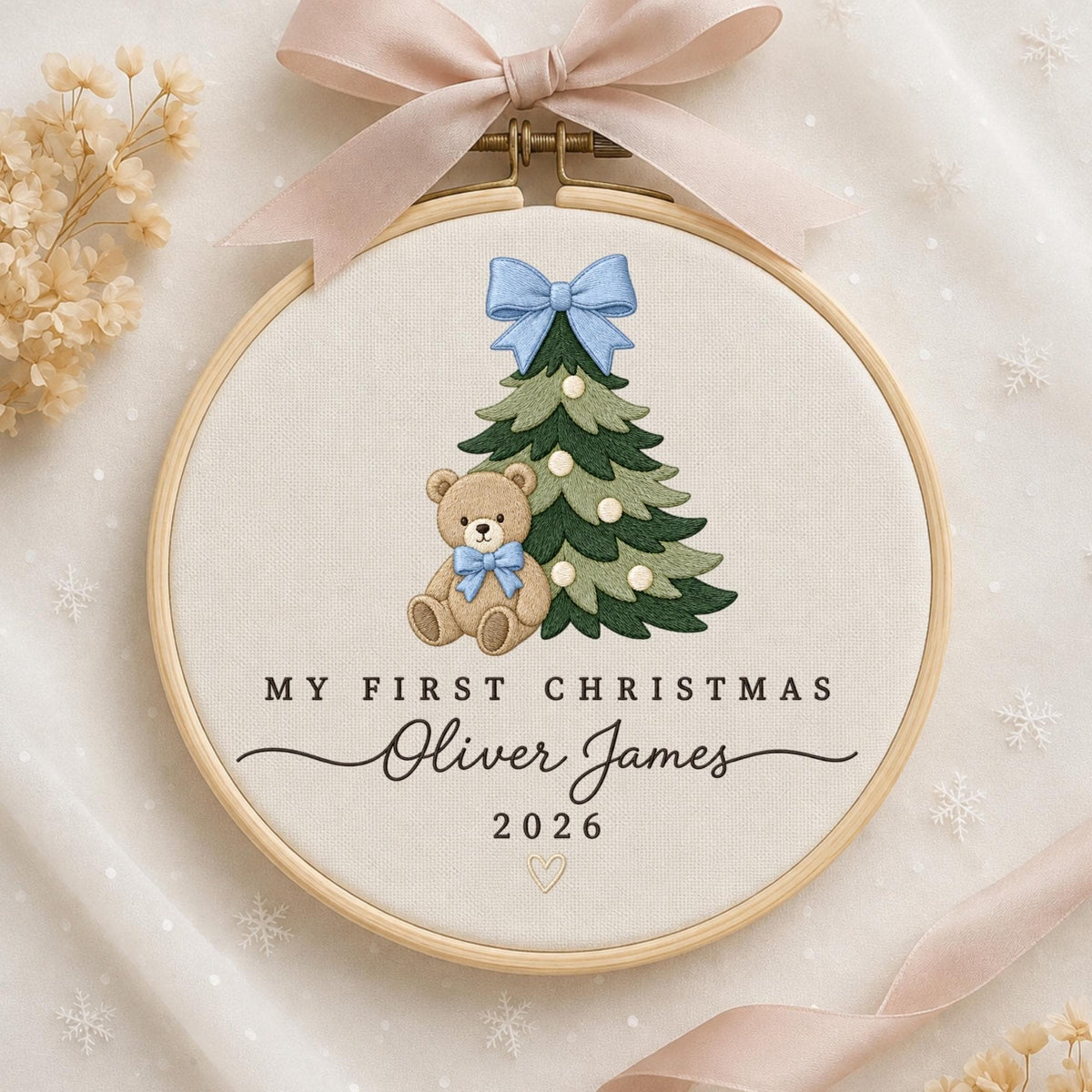 May include: A circular wooden embroidery hoop containing off-white fabric with a stitched design of a green Christmas tree and a brown teddy bear wearing a blue bow. The text reads MY FIRST CHRISTMAS, Oliver James, and 2026, with a pink ribbon.