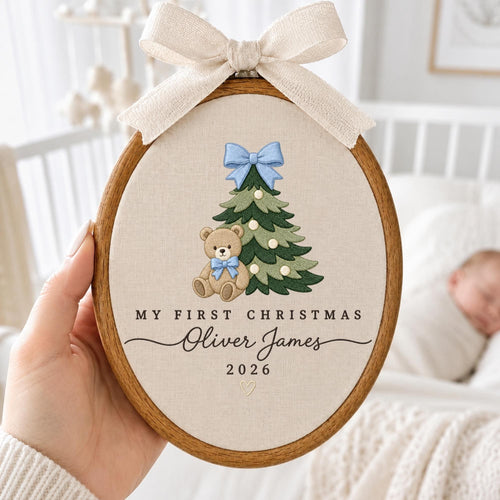 May include: An oval wooden embroidery hoop containing beige fabric with a stitched green Christmas tree, a light brown teddy bear with a blue bow, and the text "MY FIRST CHRISTMAS Oliver James 2026" with a small heart, topped with a beige ribbon.