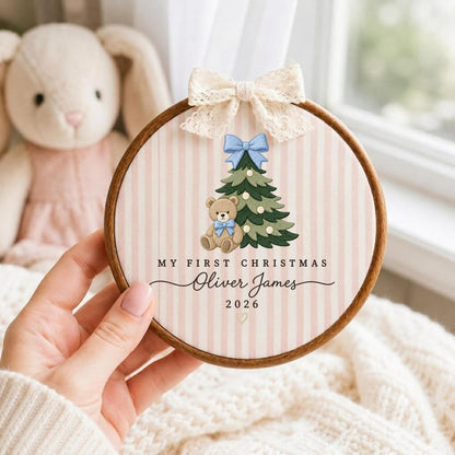 May include: A round wooden embroidery hoop featuring a light pink and white striped fabric background. The design includes an embroidered green Christmas tree with a blue bow, a teddy bear with a blue bow, and the text MY FIRST CHRISTMAS Oliver James 2026. A cream-colored lace bow is attached to the top of the hoop.