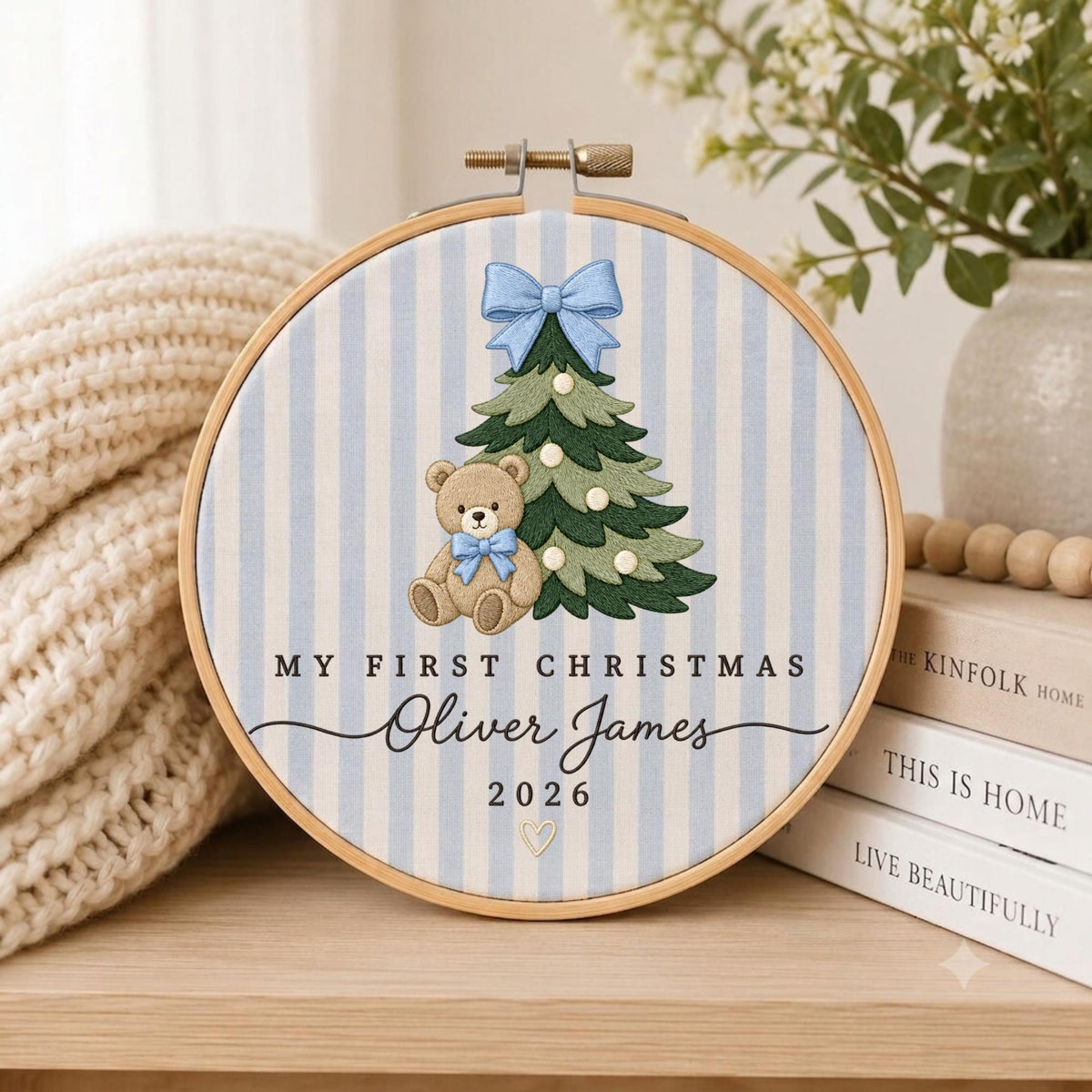 May include: A circular embroidery hoop featuring a light blue and white striped fabric background. The design includes a green Christmas tree with a light blue bow, a brown teddy bear with a blue bow, and the text My First Christmas Oliver James 2026.