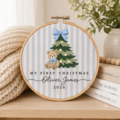 May include: A circular embroidery hoop featuring a light blue and white striped fabric background. The design includes a green Christmas tree with a light blue bow, a brown teddy bear with a blue bow, and the text My First Christmas Oliver James 2026.