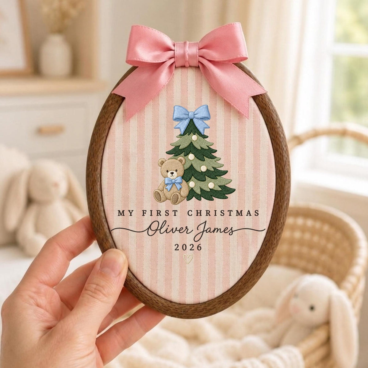 May include: An oval wooden embroidery hoop containing fabric with pink vertical stripes. It features an embroidered green Christmas tree with a light blue bow, a teddy bear with a blue bow, and the text "MY FIRST CHRISTMAS Oliver James 2026". A pink satin bow is attached to the top.