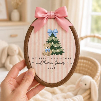 May include: An oval wooden embroidery hoop containing fabric with pink vertical stripes. It features an embroidered green Christmas tree with a light blue bow, a teddy bear with a blue bow, and the text "MY FIRST CHRISTMAS Oliver James 2026". A pink satin bow is attached to the top.