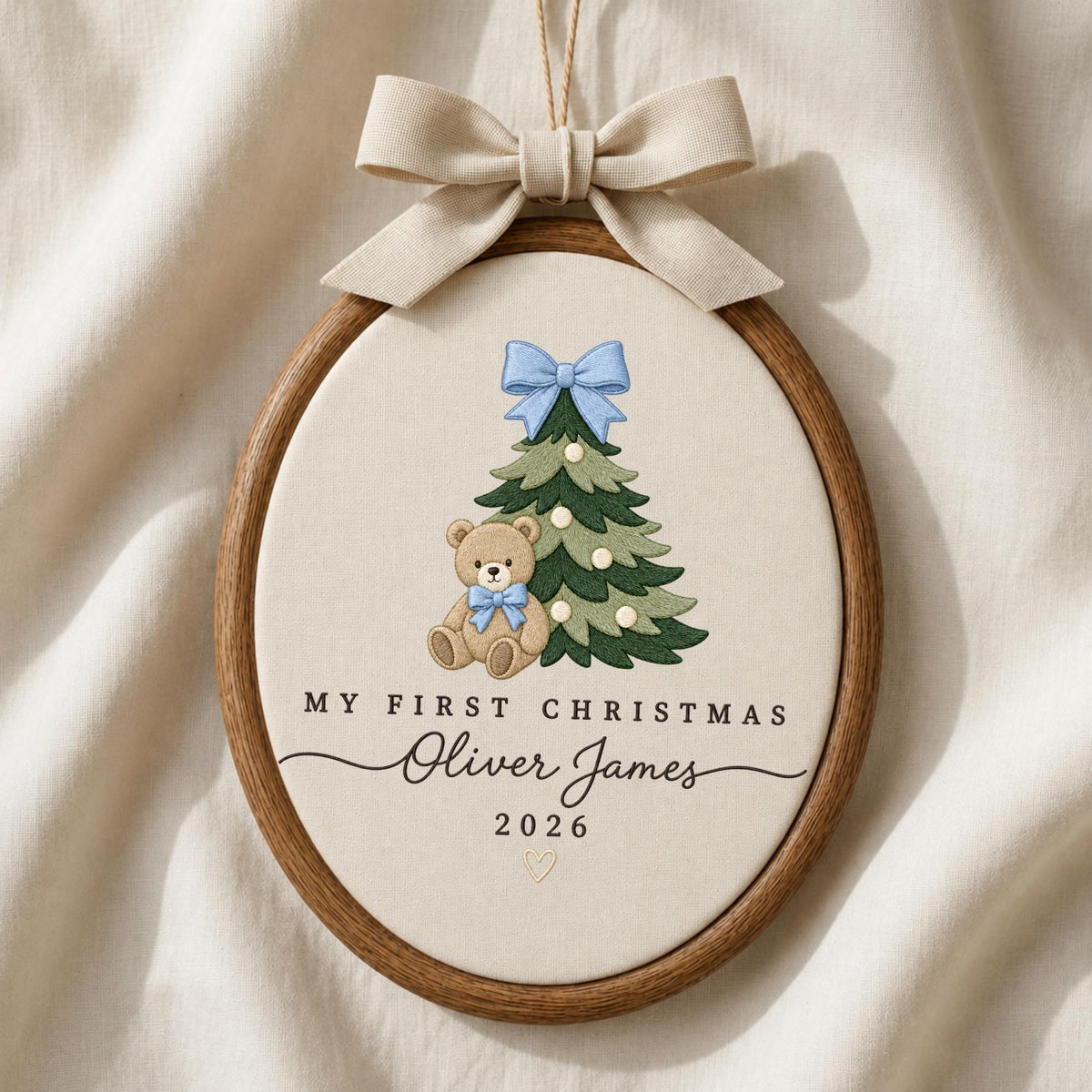 May include: An oval wooden embroidery hoop ornament featuring a beige fabric center with an embroidered green Christmas tree, a light blue bow, and a brown teddy bear. Text reads: MY FIRST CHRISTMAS, Oliver James, 2026. A beige ribbon bow hangs above.