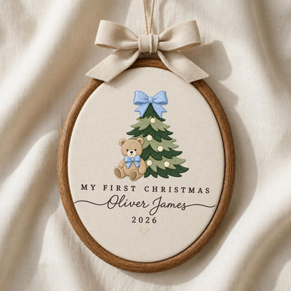 May include: An oval wooden embroidery hoop ornament featuring a beige fabric center with an embroidered green Christmas tree, a light blue bow, and a brown teddy bear. Text reads: MY FIRST CHRISTMAS, Oliver James, 2026. A beige ribbon bow hangs above.