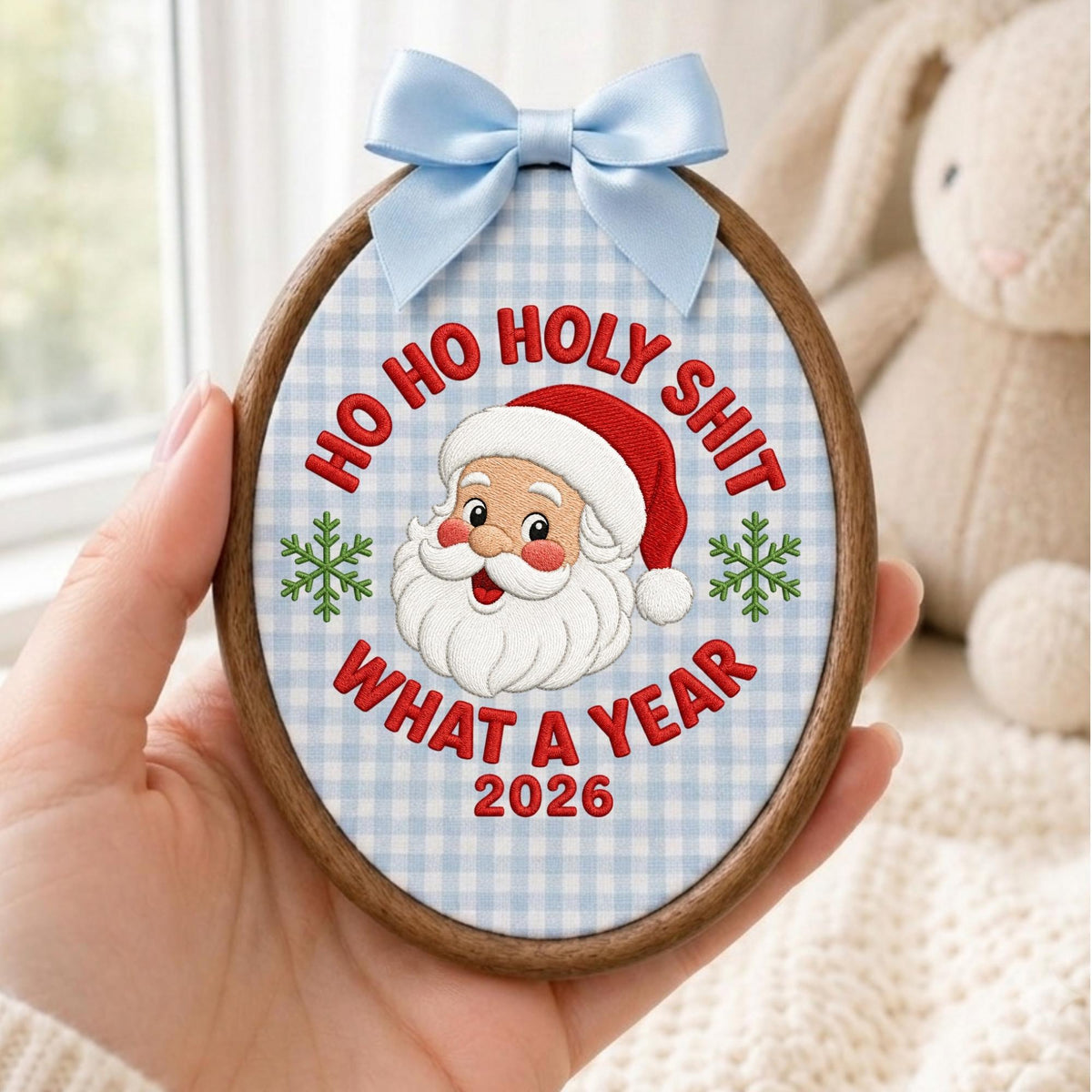 May include: An oval wooden embroidery hoop containing fabric with a light blue gingham pattern. The embroidery features a Santa Claus face, two green snowflakes, and the red text "HO HO HOLY SHIT WHAT A YEAR 2026". A light blue satin bow is attached to the top.