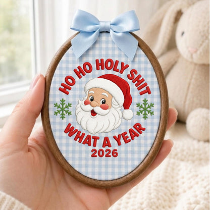 May include: An oval wooden embroidery hoop containing fabric with a light blue gingham pattern. The embroidery features a Santa Claus face, two green snowflakes, and the red text "HO HO HOLY SHIT WHAT A YEAR 2026". A light blue satin bow is attached to the top.
