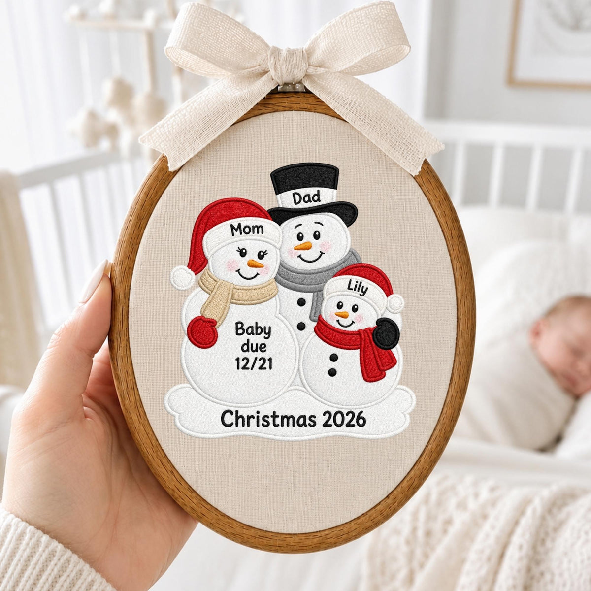 May include: An oval wooden embroidery hoop containing fabric with an embroidered design of three snowmen. The snowmen are labeled Mom, Dad, and Lily. Text on the snowmen reads Baby due 12/21, and the bottom text reads Christmas 2026. A beige ribbon bow is attached to the top.