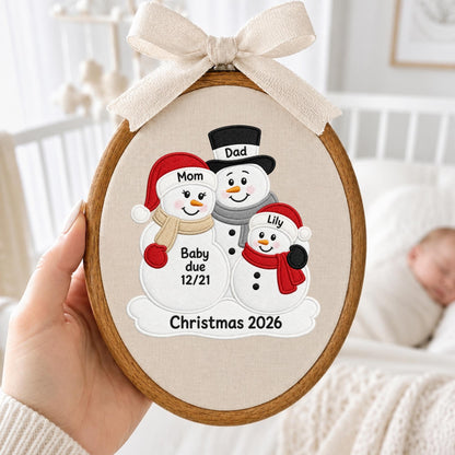 May include: An oval wooden embroidery hoop containing fabric with an embroidered design of three snowmen. The snowmen are labeled Mom, Dad, and Lily. Text on the snowmen reads Baby due 12/21, and the bottom text reads Christmas 2026. A beige ribbon bow is attached to the top.