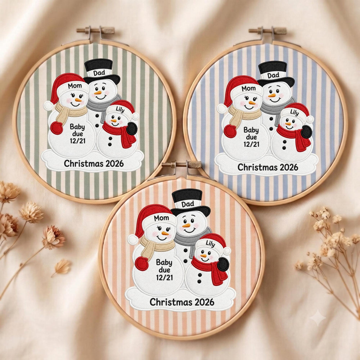 May include: Three circular embroidery hoops featuring fabric with vertical stripes in green, blue, and peach. Each hoop displays an embroidered snowman family with labels Mom, Dad, Lily, and Baby due 12/21, with the text Christmas 2026 at the bottom.