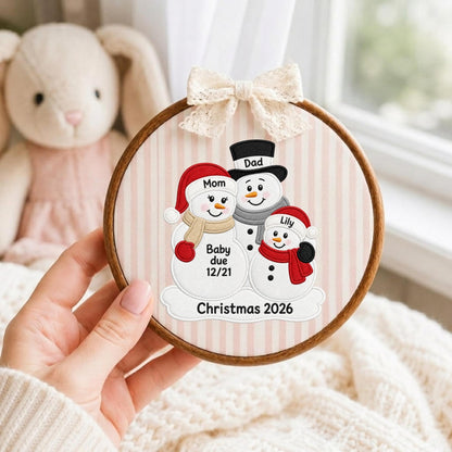 May include: A circular wooden embroidery hoop featuring a fabric background with light pink vertical stripes. The fabric displays an embroidered design of three snowmen with the text Mom, Dad, Lily, Baby due 12/21, and Christmas 2026. A cream lace bow is attached to the top.