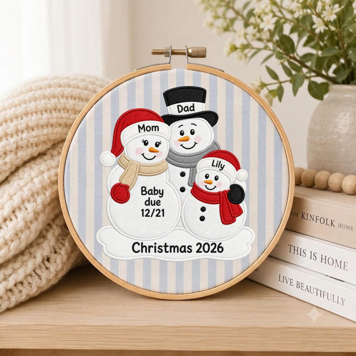 May include: A circular embroidery hoop featuring a light blue and white striped fabric background. The design displays three embroidered snowmen with the text Mom, Dad, Lily, Baby due 12/21, and Christmas 2026. The snowmen wear hats and scarves.