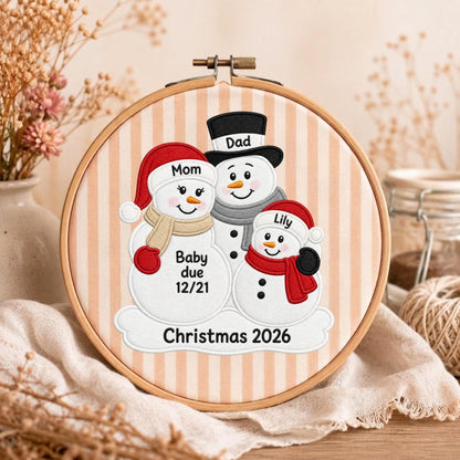 May include: A circular wooden embroidery hoop containing fabric with vertical beige stripes. The fabric features an embroidered design of three snowmen with the text Mom, Dad, Lily, Baby due 12/21, and Christmas 2026 stitched in black thread.