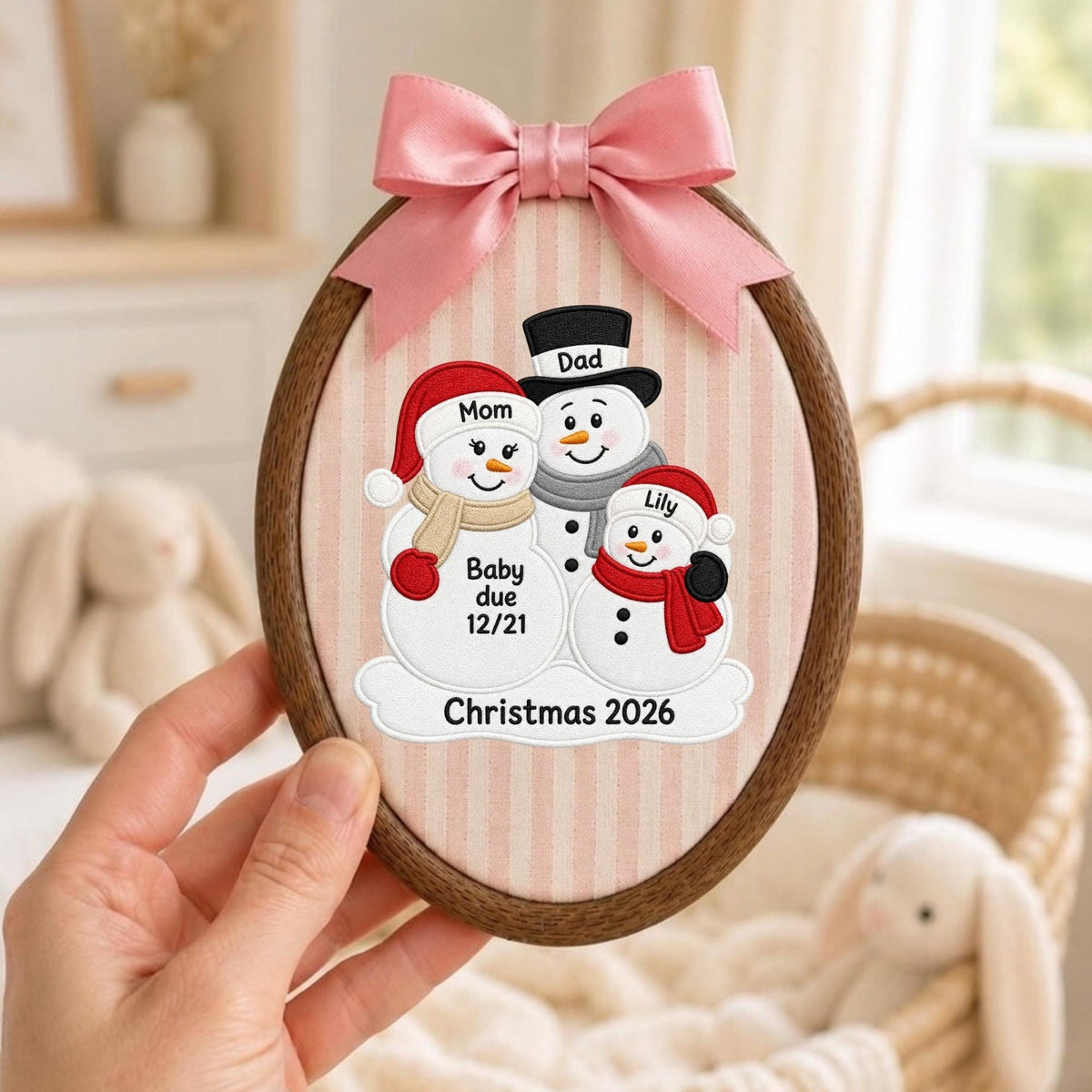 May include: An oval wooden embroidery hoop frame containing fabric with a vertical striped pattern. The fabric features an embroidered design of three snowmen labeled Mom, Dad, and Lily, with text reading Baby due 12/21 and Christmas 2026. A pink satin bow is attached to the top.