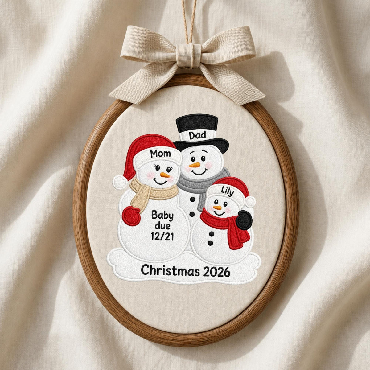May include: An oval wooden embroidery hoop containing fabric with an embroidered snowman family. The snowmen wear hats and scarves in red, tan, and grey. Text includes Mom, Dad, Lily, Baby due 12/21, and Christmas 2026. A beige ribbon bow is tied at the top.