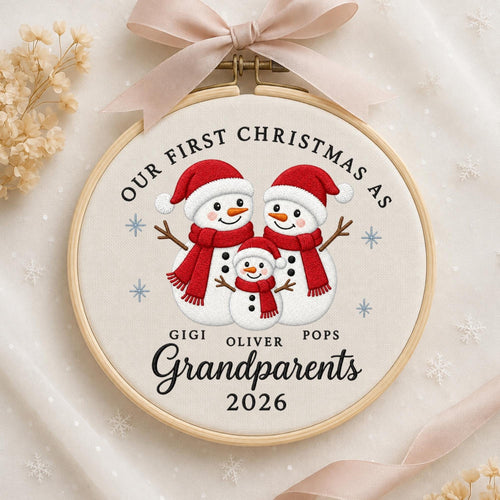 May include: A circular wooden embroidery hoop containing white fabric with an embroidered design of three snowmen wearing red hats and scarves. The text reads OUR FIRST CHRISTMAS AS GIGI OLIVER POPS Grandparents 2026. A beige ribbon is tied at the top.
