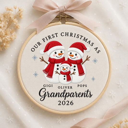 May include: A circular wooden embroidery hoop containing white fabric with an embroidered design of three snowmen wearing red hats and scarves. The text reads OUR FIRST CHRISTMAS AS GIGI OLIVER POPS Grandparents 2026. A beige ribbon is tied at the top.
