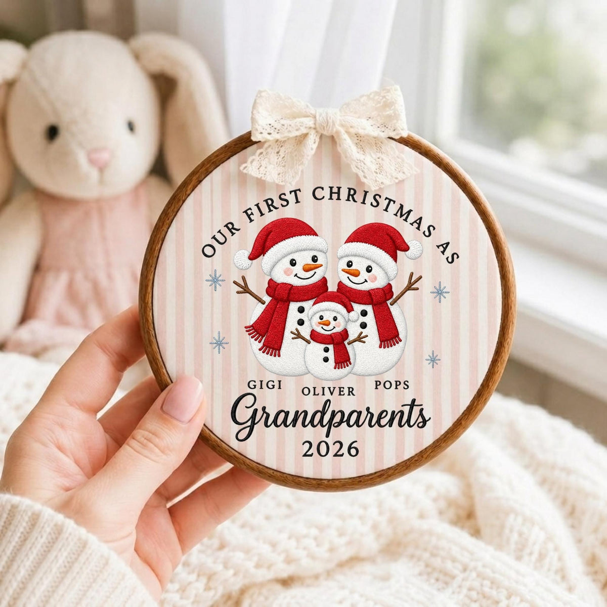 May include: A round wooden embroidery hoop ornament featuring a light pink and white striped background. It displays three illustrated snowmen wearing red hats and scarves, with the text "OUR FIRST CHRISTMAS AS GIGI OLIVER POPS Grandparents 2026".