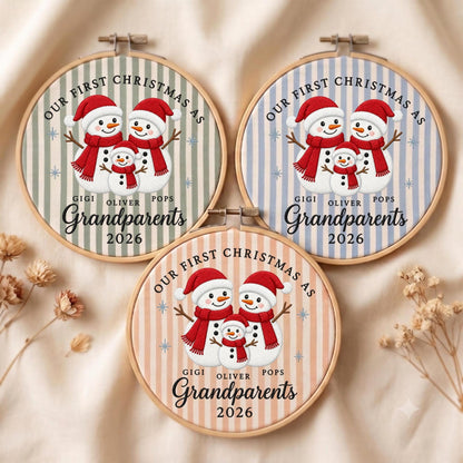 May include: Three circular wooden embroidery hoops featuring fabric with vertical stripes in sage green, light blue, and peach. Each displays three snowmen wearing red hats and scarves, with the text "OUR FIRST CHRISTMAS AS GIGI OLIVER POPS Grandparents 2026".