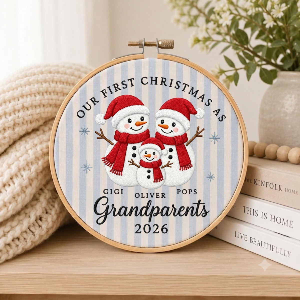 May include: A circular embroidery hoop featuring a light blue and white striped fabric background. The design displays three snowmen wearing red hats and scarves, with the text "OUR FIRST CHRISTMAS AS GIGI OLIVER POPS GRANDPARENTS 2026".