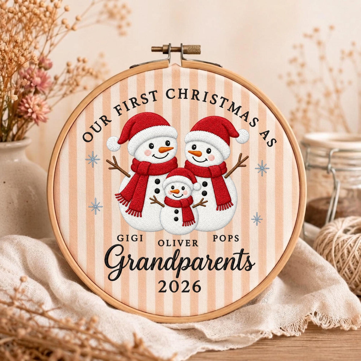 May include: An embroidery hoop featuring a printed design of three snowmen wearing red hats and scarves. The text reads: OUR FIRST CHRISTMAS AS GIGI OLIVER POPS Grandparents 2026. The background has vertical beige stripes and blue snowflake accents.